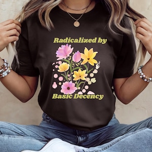 Peut inclure: T-shirt noir avec un motif floral composé de fleurs roses et jaunes et le texte "Radicalized by Basic Decency" en jaune. Le t-shirt est en matière douce, idéal pour un usage quotidien.