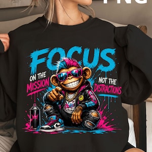 Peut inclure: Sweat-shirt noir avec un singe souriant portant des lunettes de soleil et une veste. Le texte "FOCUS ON THE MISSION NOT THE DISTRACTIONS" est en bleu et rose. Un aérosol est à gauche. Style graffiti.
