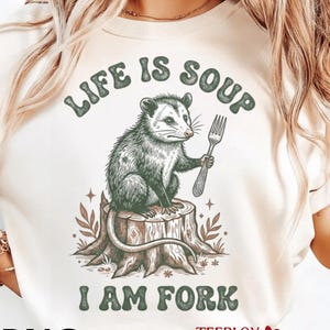 Peut inclure: T-shirt crème avec un motif graphique d'un opossum tenant une fourchette, assis sur une souche d'arbre. Le texte "LIFE IS SOUP I AM FORK" est au-dessus et en dessous de l'opossum. Le design comprend des éléments botaniques et des étoiles.