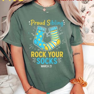 Peut inclure: T-shirt vert avec l'inscription "Proud Sibling" et "Rock Your Socks" en jaune et bleu. Le motif comprend deux chaussettes, une bleue à pois jaunes et l'autre jaune à rayures bleues. La date "21 mars" est également imprimée.