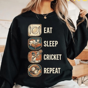 Peut inclure: Sweat-shirt noir avec l'inscription blanche "EAT SLEEP CRICKET REPEAT". Le motif comprend des illustrations d'une assiette et de couverts, d'un lit avec un livre et un réveil, d'un joueur de cricket et d'une horloge.