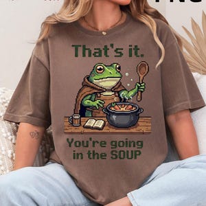 Peut inclure: T-shirt marron avec une illustration de chef grenouille en pixel art. La grenouille, portant un chapeau et un tablier de chef, remue une marmite de soupe. Le texte sur le t-shirt se lit "That's it. You're going in the SOUP."
