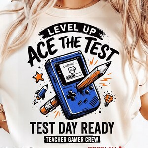 Peut inclure: Un t-shirt crème avec un motif graphique représentant une console de jeu portable bleue avec un crayon qui la traverse. Le texte indique "LEVEL UP ACE THE TEST" et "TEST DAY READY TEACHER GAMER CREW".