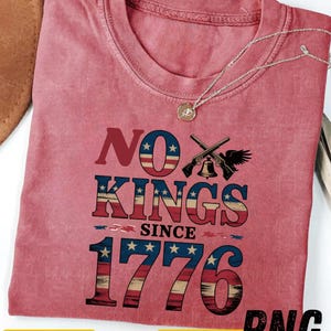 Peut inclure: Un t-shirt rouge avec le texte "NO KINGS SINCE 1776" dans un design patriotique. Le motif comprend des fusils croisés, une cloche et un aigle. Un collier argenté avec un pendentif est drapé sur le t-shirt.