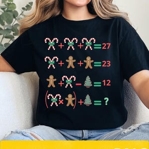 Peut inclure: T-shirt noir avec une équation de Noël utilisant des cannes en sucre, des bonhommes en pain d'épices et des sapins. Le texte "DIGITAL PRODUCTS PNG" est également inclus.
