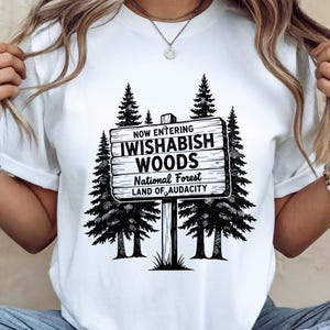 Peut inclure: T-shirt blanc avec un motif noir et blanc d'un panneau en bois indiquant "NOW ENTERING IWISHABISH WOODS National Forest LAND OF AUDACITY" et des pins. Le t-shirt a un col rond.
