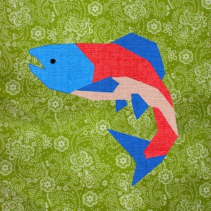 Op de afbeelding: Een rode, blauwe en roze vis-applicatie op een groene stof met een wit bloemenpatroon.
