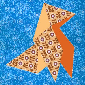 Può includere: Un uccello di origami colorato fatto di tessuto con un motivo floreale marrone e bianco, tessuto arancione e pesca, su uno sfondo blu con un motivo floreale bianco.