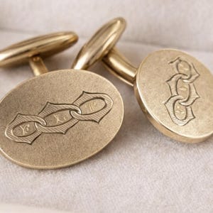 May include: A pair of oval-shaped gold-toned cufflinks featuring a matte finish and an engraved design of three interlocking chain links. Each cufflink has a smooth, rounded face with a central motif and a standard post and toggle mechanism.
