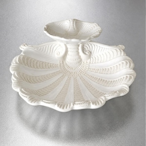 May include: A white ceramic two-tiered serving dish shaped like a large scallop shell. The base is a wide, fluted shell with intricate radial ridges and a textured surface, supporting a smaller, matching shell-shaped bowl on a central pedestal.