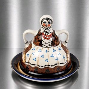 May include: A ceramic butter dish featuring a lid shaped like a person wearing a white headscarf, a brown bodice with red beaded necklace detail, and a white skirt with blue floral accents. The lid has two handles and sits on a matching plate.