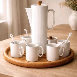 Puede incluir: Un juego de café de cerámica blanca que incluye una cafetera alta y acanalada con un pomo de madera en la tapa, cuatro tazas acanaladas a juego y cuatro cucharas blancas. El conjunto está dispuesto sobre una bandeja redonda de madera.