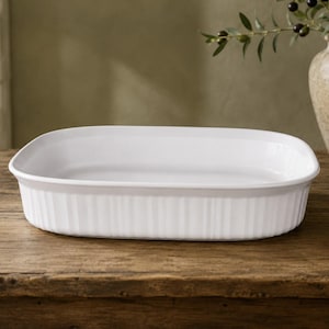 May include: A rectangular white ceramic baking dish with rounded corners and a fluted exterior design. The dish features a smooth interior surface and a thick, sturdy rim, providing a clean and functional appearance for oven-to-table food preparation.