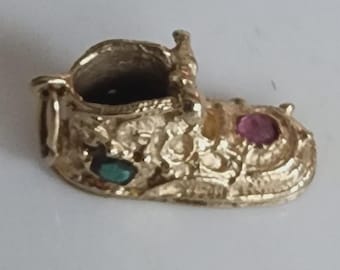 Op de afbeelding: Een goudkleurige babyschoenbedel met getextureerde details. De bedel heeft een kleine, open bovenkant en is versierd met een groene en een roze edelsteen. Dit sieradenartikel is waarschijnlijk een hanger of bedel.