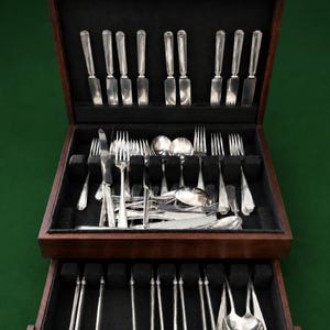 May include: A wooden flatware chest with a hinged lid and a pull-out drawer, featuring a black velvet interior. The set includes silver-toned knives, forks, and spoons arranged in designated slots, with additional pieces stored in the lower drawer.