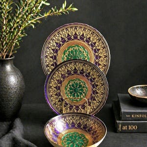 May include: Two circular plates and one matching bowl featuring a deep purple and emerald green color palette with intricate gold filigree patterns. The plates are displayed vertically on a stand, while the bowl sits in front on a dark surface.