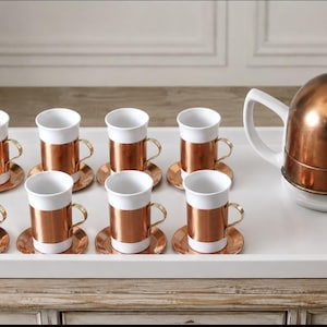May include: A tea set consisting of eight white ceramic cups with copper sleeves and matching saucers, accompanied by a white ceramic teapot with a copper dome lid, all arranged on a large white rectangular serving tray with side handles.