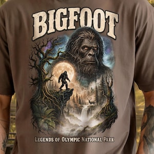 Può includere: Una t-shirt marrone con una stampa grafica che raffigura un grande volto di Bigfoot sopra una sagoma più piccola di Bigfoot in piedi su una scogliera. Il design include una luna piena, una foresta, una cascata e il testo BIGFOOT Legends of Olympic National Park.