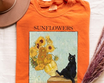 May include: Orange crew neck t-shirt featuring a painting of sunflowers in a vase and a black cat. The word "SUNFLOWERS" is printed above the painting. The shirt is folded on a white surface.