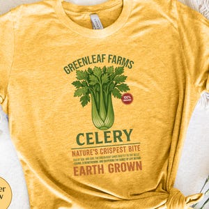 May include: Heather yellow gold t-shirt featuring a celery graphic. The design includes the text "GREENLEAF FARMS", "CELERY", and "NATURE'S CRISPEST BITE". The shirt also has the text "EARTH GROWN" and a "100% organic" label.