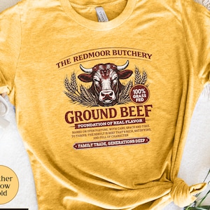 May include: Heather yellow gold t-shirt with a graphic for "The Redmoor Butchery", a bull illustration, and the words "Ground Beef". The shirt also features the text "100% Grass Fed" and "Family Trade, Generations Deep".