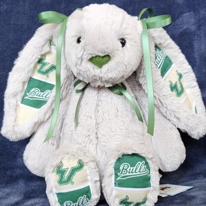 May include: A plush stuffed rabbit with soft light gray fur, featuring long ears and feet accented with green and cream fabric panels. The fabric displays the green USF Bulls logo and the word Bulls in white text. A green satin ribbon is tied at the neck.