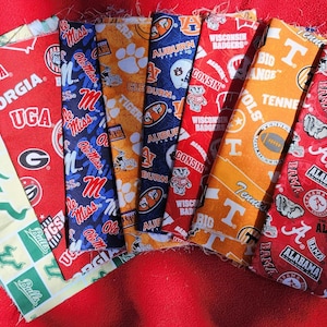 May include: A collection of seven rectangular fabric swatches featuring various collegiate sports team logos and colors, including green, red, blue, orange, and white patterns with printed text such as Georgia, Ole Miss, Auburn, Wisconsin, and Alabama.