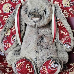 May include: A plush stuffed rabbit with mottled gray and brown fur, featuring long ears lined with red fabric printed with Florida State University logos and text. The rabbit has a pink nose, black eyes, and two cream and red ribbons tied at the top.