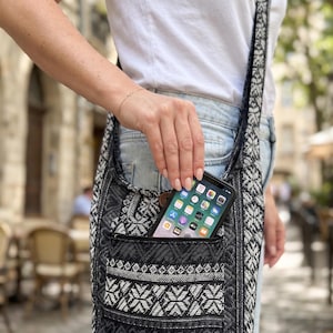 May include: A black and white woven crossbody bag featuring a repeating geometric snowflake and heart pattern. The bag has a wide shoulder strap and an external front pocket with a zipper closure, currently holding a black smartphone.