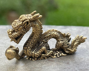 May include: A small, gold-colored dragon figurine with intricate details. The dragon is curled up with its tail wrapped around its body, and it is holding a pearl in its mouth.