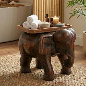May include: A carved wooden elephant stool with a tray holding rolled white towels, small bottles, and a candle. The stool is brown and sits on a woven rug. The scene suggests a spa or bathroom setting.
