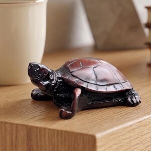 May include: A light-colored wooden floating shelf holding a cream-colored ceramic mug, a small dark-brown turtle figurine with a textured shell, a gray geometric planter with a green succulent, and a stack of three brown leather-bound books.