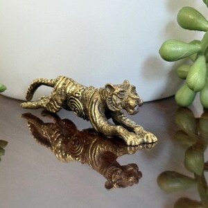 Op de afbeelding: Een gouden tijgerfiguur in kruipende houding, met gedetailleerde ge&euml;tste patronen op het lichaam. De figuur wordt weerspiegeld op een glanzend oppervlak. Dit decoratieve item kan een verzamelobject of een stukje woondecoratie zijn.