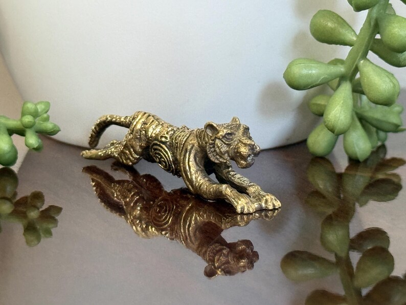 Op de afbeelding: Een gouden tijgerfiguur in kruipende houding, met gedetailleerde ge&euml;tste patronen op het lichaam. De figuur wordt weerspiegeld op een glanzend oppervlak. Dit decoratieve item kan een verzamelobject of een stukje woondecoratie zijn.