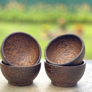 May include: Four small, round wooden bowls stacked in pairs. The bowls are a dark brown color with a speckled pattern. The background is a blurred green and gray, suggesting an outdoor setting.