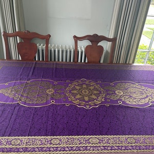 May include: Purple tablecloth with a gold embroidered design featuring floral patterns and a central mandala. The tablecloth is laid on a wooden table with two chairs.