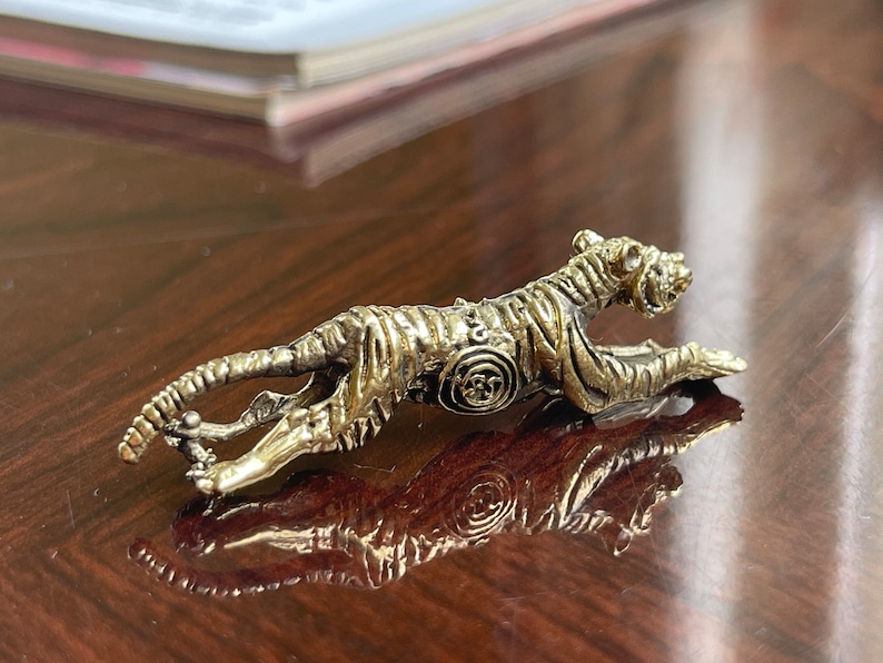 Op de afbeelding: Gedetailleerd metalen tijgerbeeldje in een rennende pose. De tijger is goud- en zilverkleurig, met ingewikkelde details op zijn lichaam. Het beeldje is ongeveer 10 cm lang en wordt getoond op een reflecterend oppervlak.