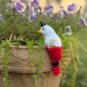 May include: A decorative ceramic bird figurine with a white body, red tail, and black and yellow accents. The bird is perched on the edge of a brown flower pot filled with green plants and purple flowers.