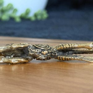 Op de afbeelding: Een gedetailleerd, goudkleurig beeldje van een mythisch wezen, mogelijk een draak of een soortgelijk wezen. De sculptuur ligt, met ingewikkelde details zichtbaar op het lichaam, inclusief schubben en sierlijke patronen. Het beeldje staat op een houten oppervlak.
