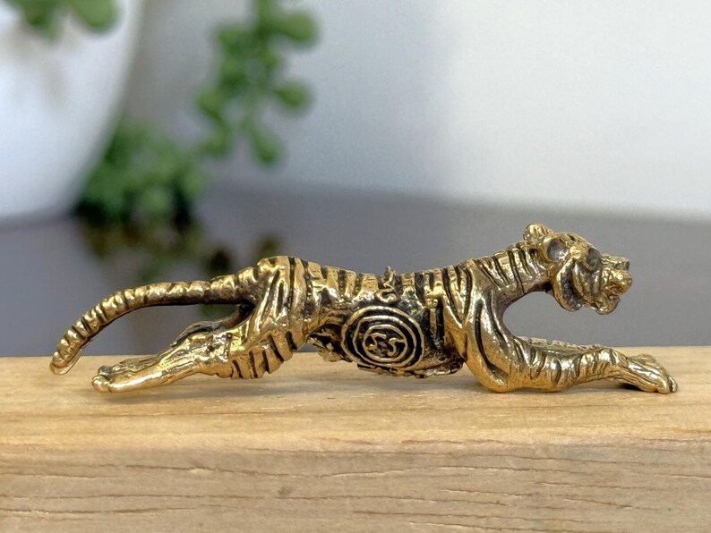 Op de afbeelding: Een messing tijgerfiguur in een hurkende houding, met gedetailleerde strepen en een cirkelvormig ontwerp aan de zijkant. De goudkleurige sculptuur staat op een houten oppervlak, wat het vakmanschap benadrukt. Ongeveer 10 cm lang.