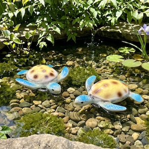 May include: Two ceramic turtle figurines with yellow and brown patterned shells and blue flippers float in a pond. The pond is surrounded by rocks, greenery, and white flowers. The turtles are a decorative garden accent.