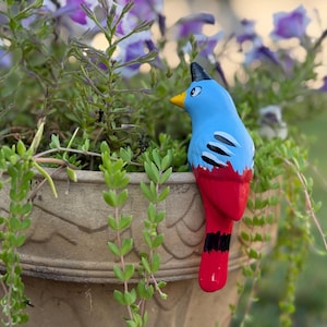 May include: A ceramic bird figurine with a blue body, red tail, and black and white accents. The bird has a yellow beak and a black crest. It is placed near a flower pot filled with green plants and purple flowers.
