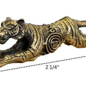 Op de afbeelding: Een bronzen tijgerbeeldje in een springhouding. De sculptuur is 5,7 cm lang, 1,9 cm breed en 1,3 cm hoog. De tijger heeft gedetailleerde strepen en gezichtskenmerken, met een cirkelvormig ontwerp aan de zijkant.