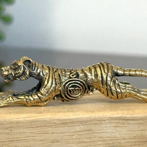 Op de afbeelding: Gedetailleerd messing tijgerbeeldje in een rennende pose. De tijger heeft zwarte strepen en ligt op een houten oppervlak. Het beeldje is een decoratief item.