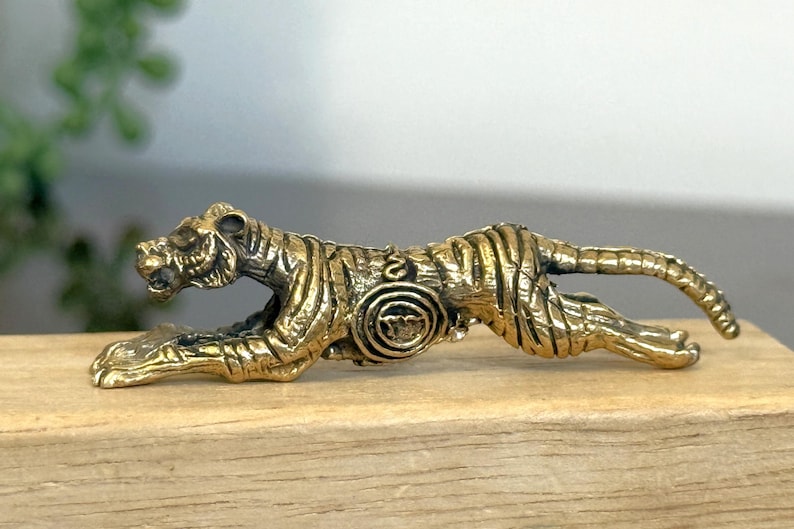 Op de afbeelding: Gedetailleerd messing tijgerbeeldje in een rennende pose. De tijger heeft zwarte strepen en ligt op een houten oppervlak. Het beeldje is een decoratief item.