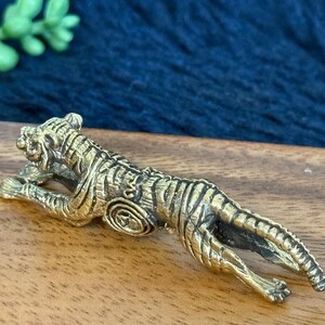 Op de afbeelding: Een gedetailleerd messing tijgerbeeldje, liggend met zijn kop omhoog. De tijger heeft ingewikkelde gestreepte details en is geplaatst op een houten oppervlak. Het beeldje is een decoratief item.