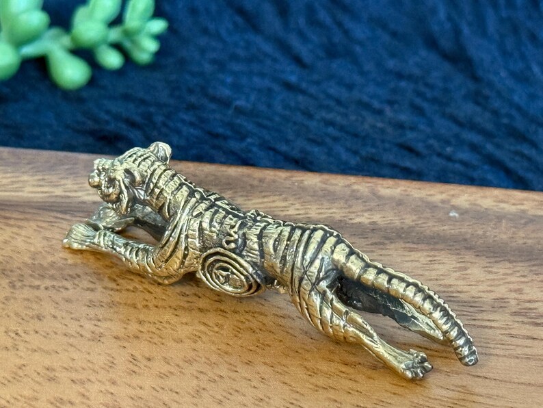 Op de afbeelding: Een gedetailleerd messing tijgerbeeldje, liggend met zijn kop omhoog. De tijger heeft ingewikkelde gestreepte details en is geplaatst op een houten oppervlak. Het beeldje is een decoratief item.