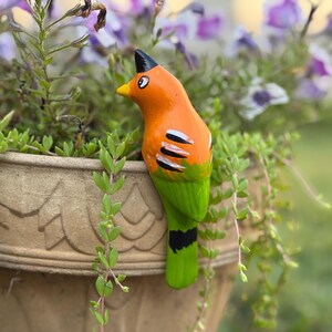 May include: A vibrant, hand-painted bird figurine with an orange body, green tail, and black accents. The bird has a yellow beak and a black crest. It is perched on the edge of a decorative flower pot filled with greenery and purple flowers.
