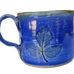 May include: A handmade ceramic mug with a deep blue glaze. The mug features a leaf imprint on the side and a textured handle. The rim has a contrasting green-blue hue. The mug is suitable for hot or cold beverages.