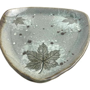 May include: A ceramic, irregularly shaped plate with a gray-green background. The plate features a leaf design in gray and brown, with additional brown and white speckles. The rim of the plate has a darker, brownish color.